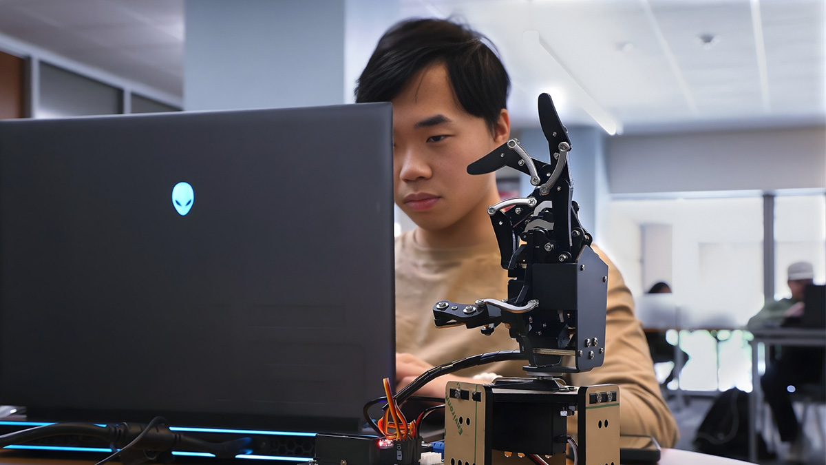  A student sitting in a library, with a laptop in front of him and mechanical hand beside him, programming its movements